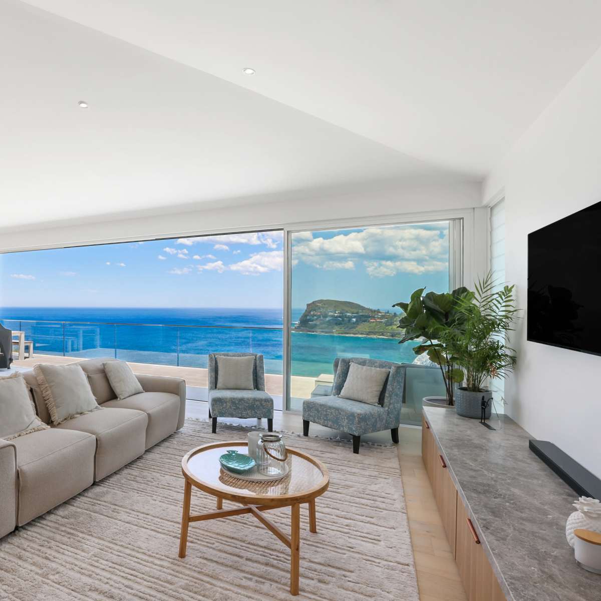 A modern living room features a large window with a view of the ocean, furnished with a beige sofa, a wooden coffee table, and a dining area in the background.