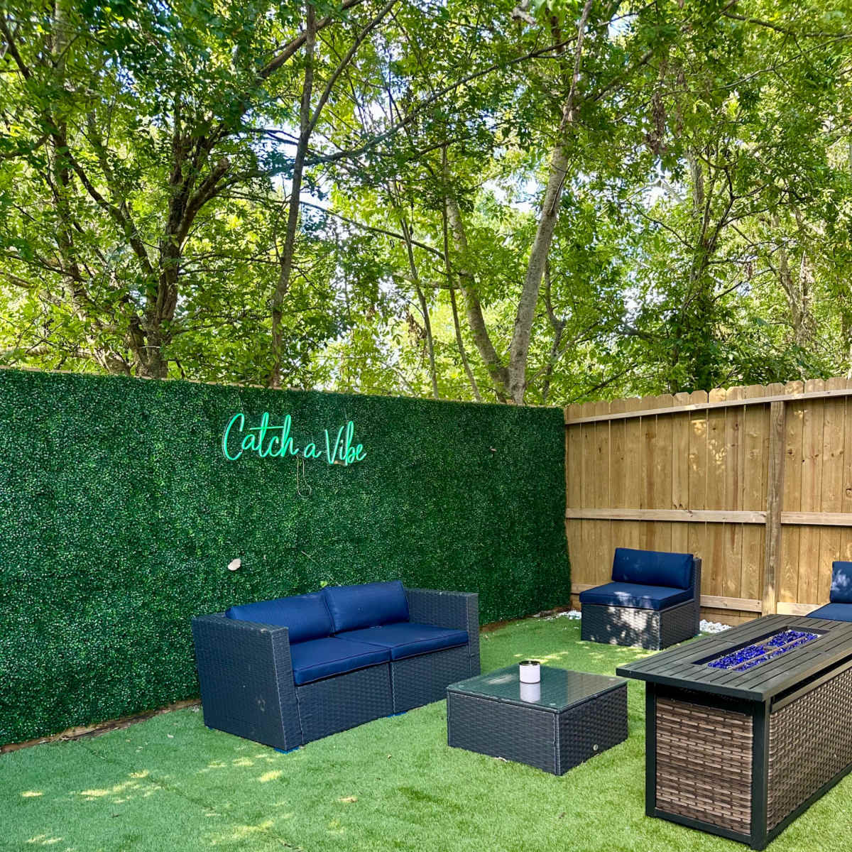 A landscaped outdoor space with synthetic grass, seating couches, a coffee table, and a fire pit against a green wall.