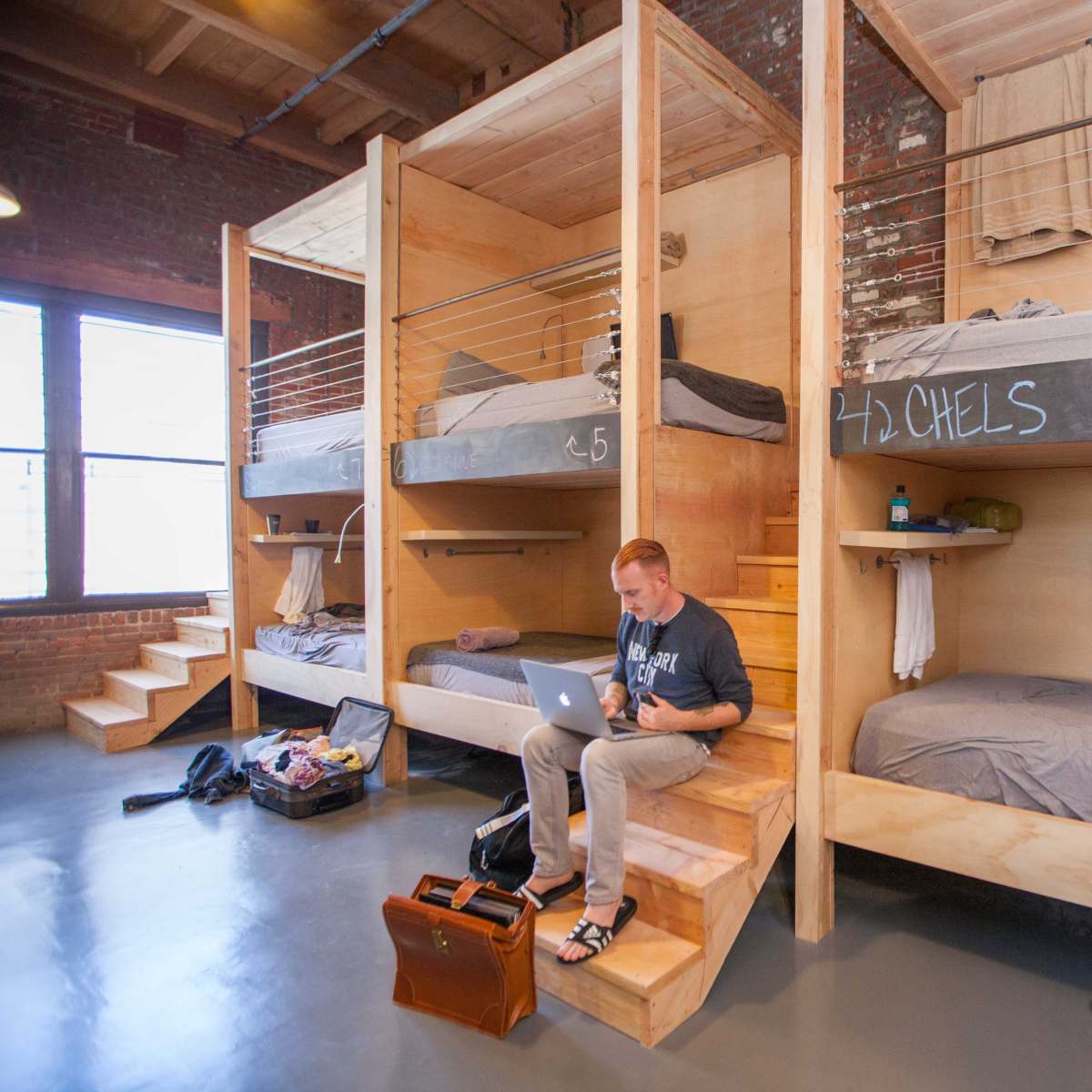 A modern dormitory-style room with wooden bunk beds and a man sitting on the stairs using a laptop.