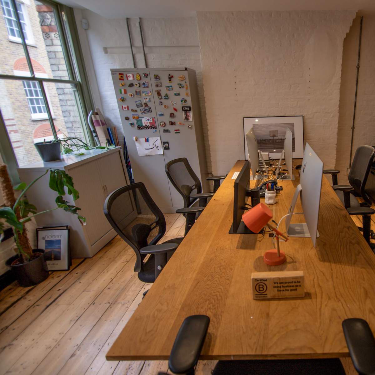 A spacious office features a long wooden table surrounded by black chairs, with a computer and lamp on the table, and a fridge covered in magnets in the background.