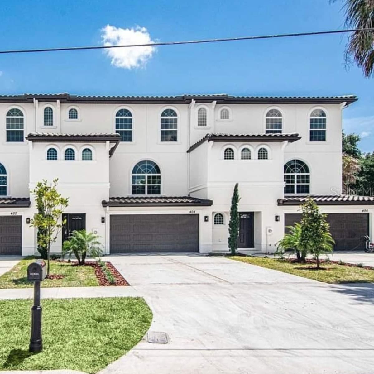 A row of three modern white multi-family homes with arched windows and a paved driveway, all set against a clear blue sky.