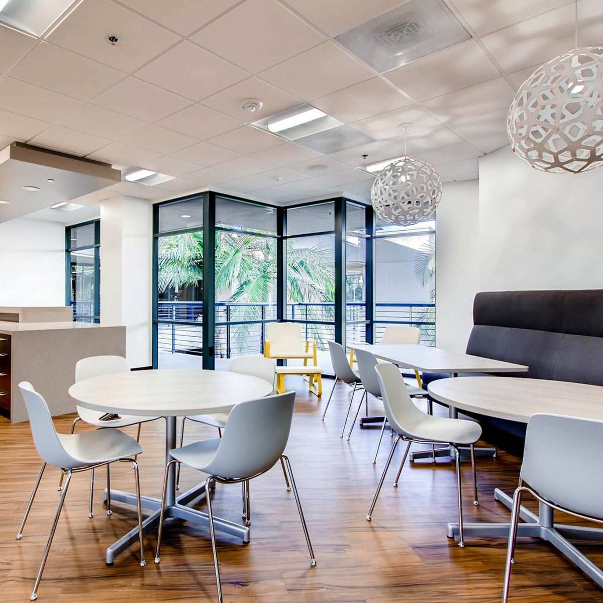 A modern kitchen or break area with light wood flooring, round tables, white chairs, and large windows overlooking greenery.