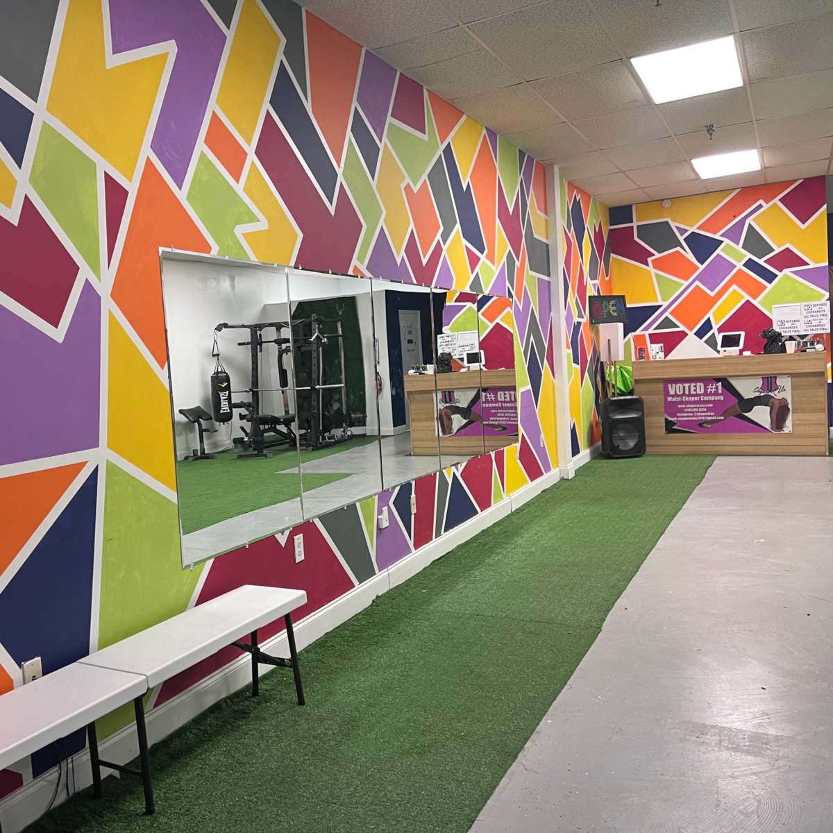A brightly colored gym interior with a geometric mural on the walls, a mirror, a bench, and a patch of green turf.