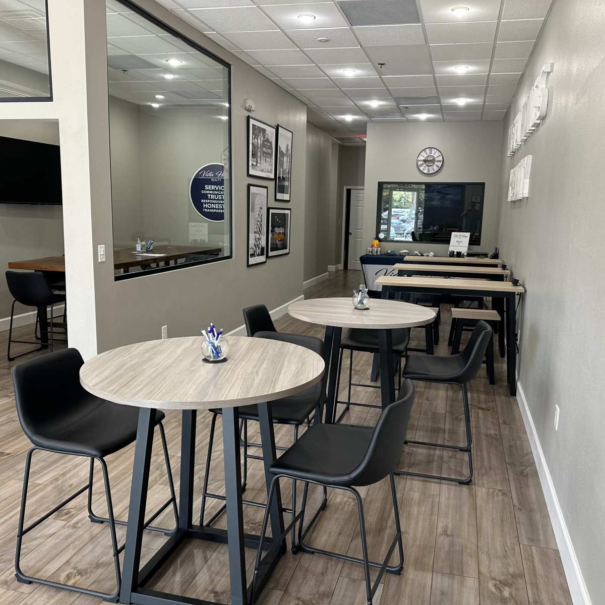 A modern, minimalistic interior of a cafe or office space featuring small round tables and black chairs, with a large window and various wall decorations.