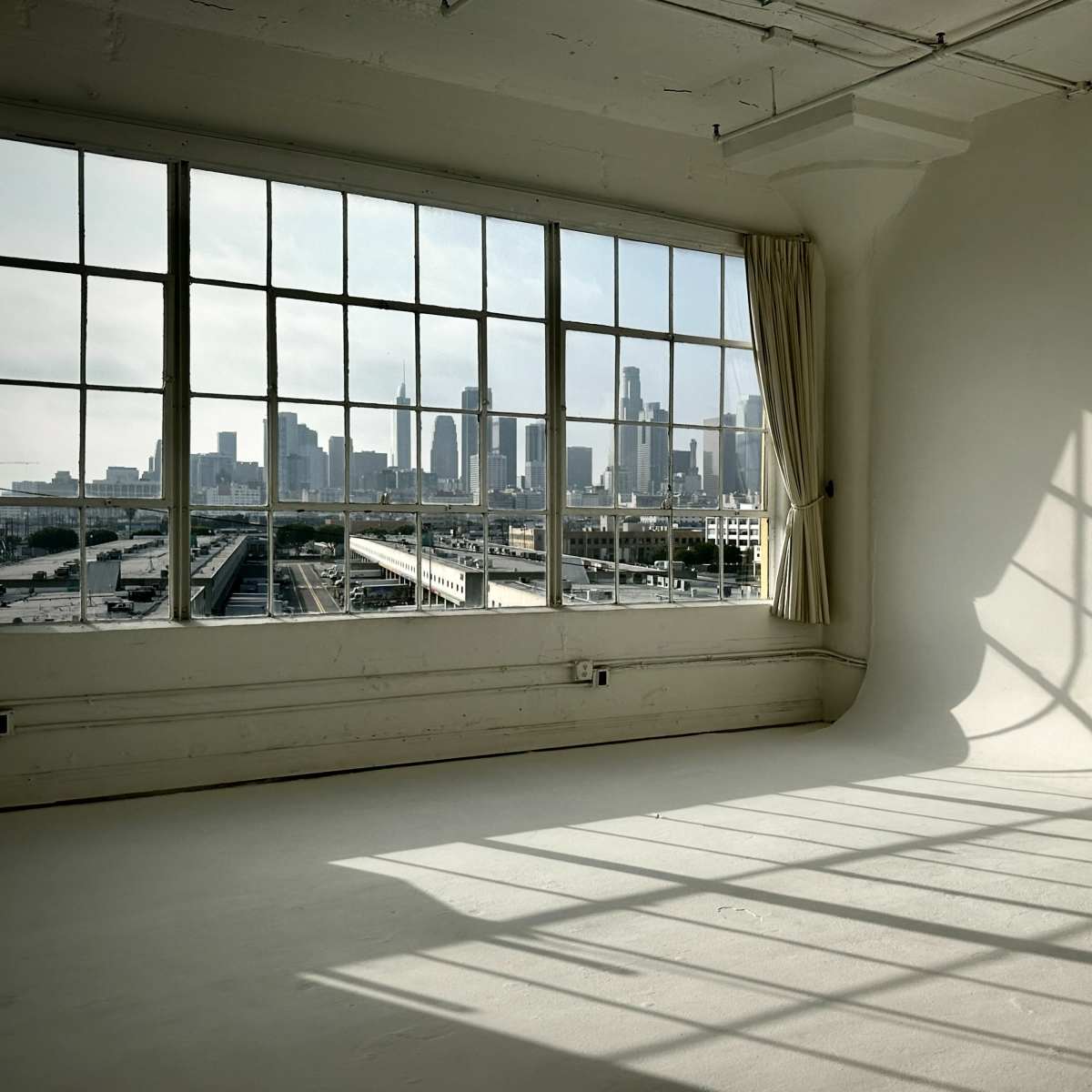 An empty room with large windows offering a view of a city skyline, casting shadows on the floor.
