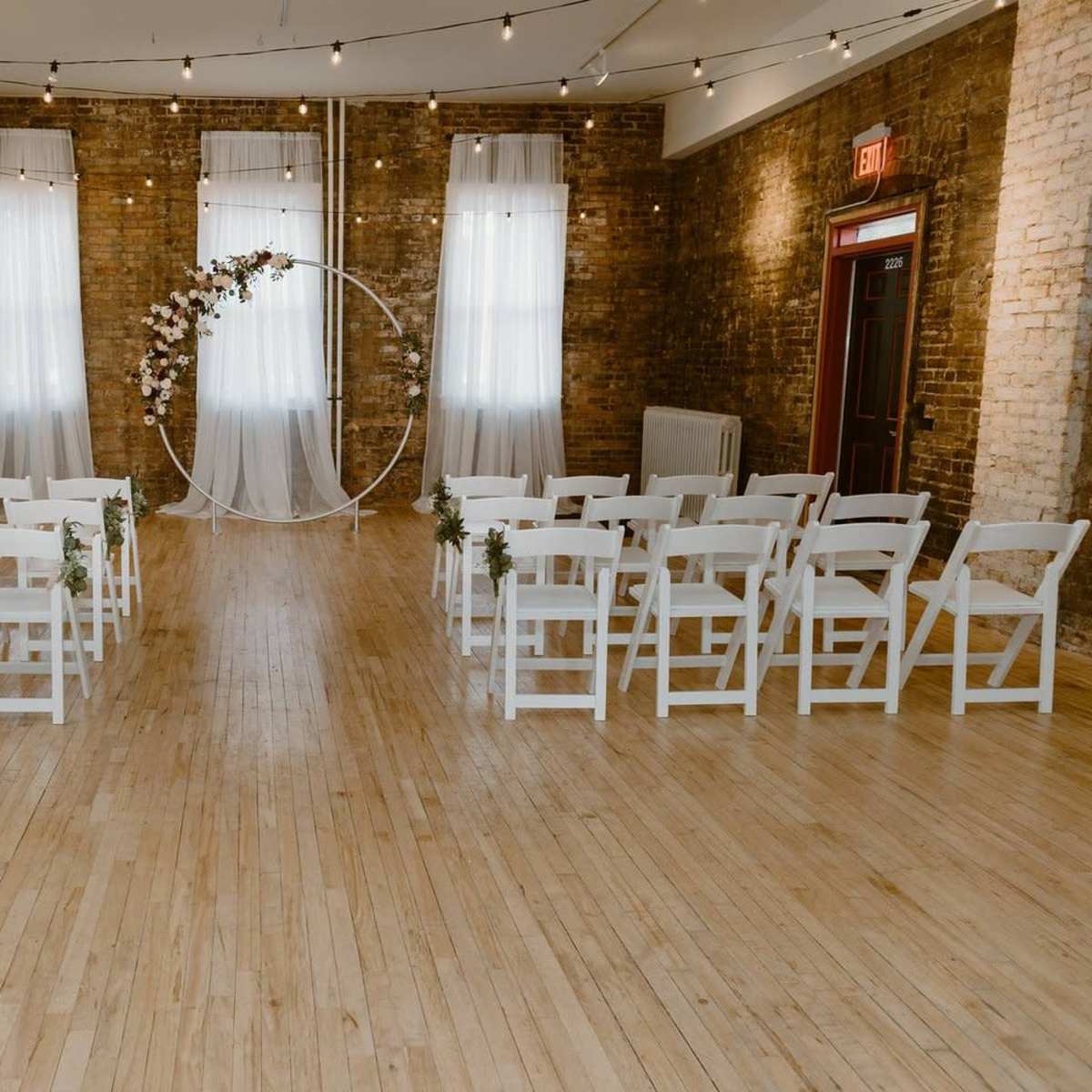 A spacious venue features neatly arranged white chairs facing a circular floral arch, with large windows and exposed brick walls in the background.