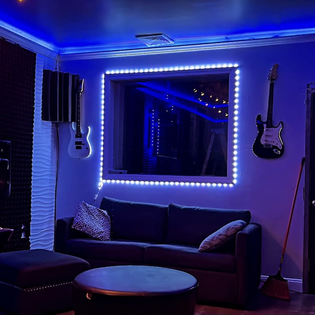 A music studio room with a dark sofa, a circular table, guitar hangers on the wall, soundproofing panels, and blue LED lighting around a window.