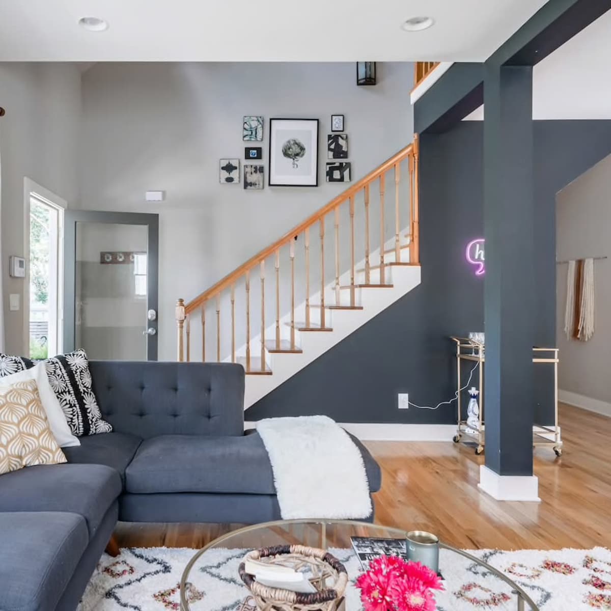 A modern living room features a sectional couch, a round glass coffee table, and a staircase adorned with framed artwork.