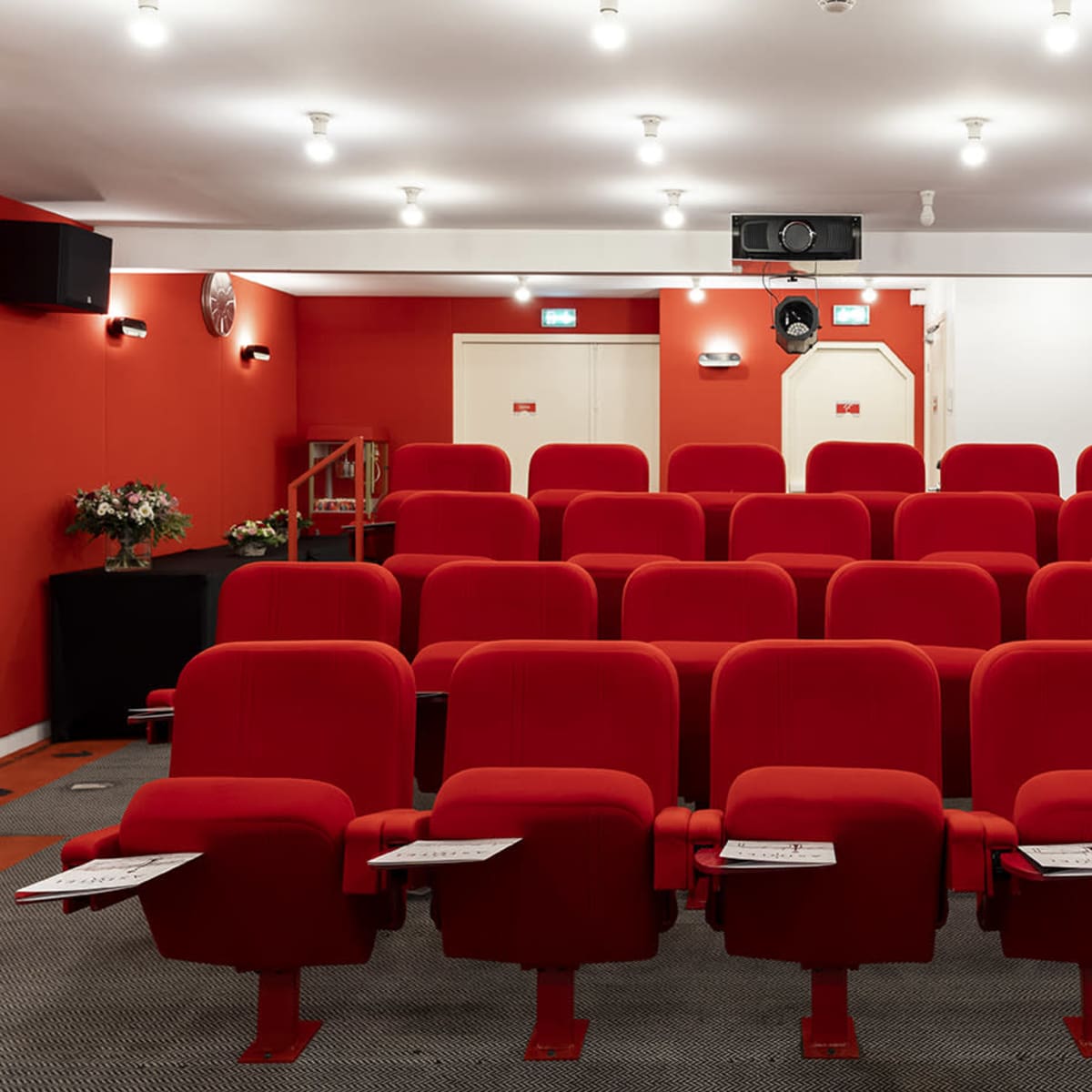 A small theater features red upholstered seats arranged in rows facing a projector screen, with a simple interior design.