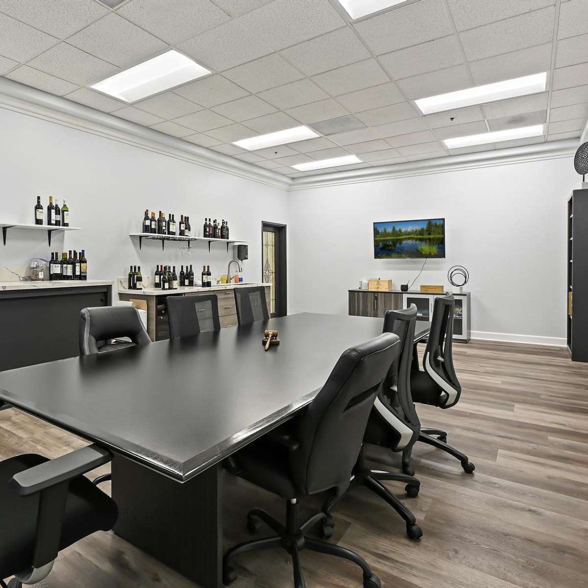 A modern conference room with a large black table, black ergonomic chairs, and shelves lined with various bottles, complemented by a television on the wall.