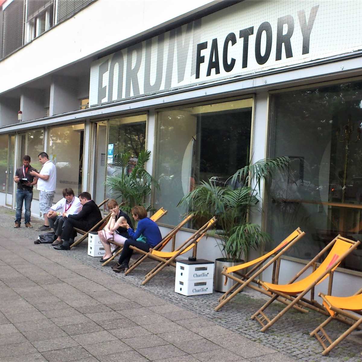 A group of people is seated on deck chairs outside a building labeled "FIRMIT Factory," with large windows and potted plants.