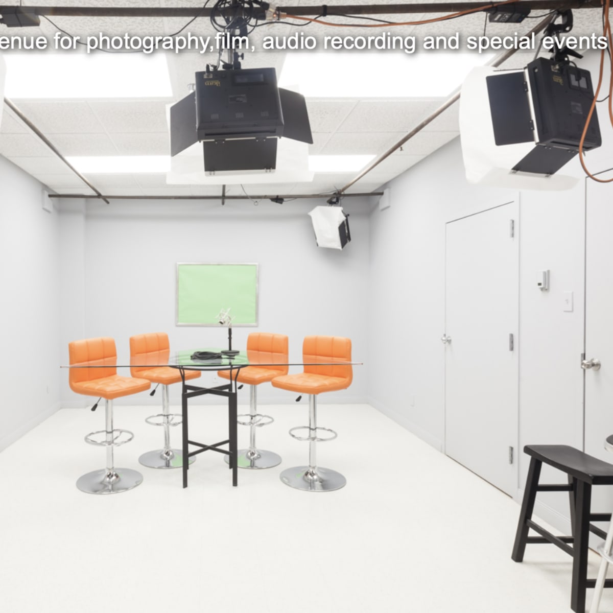 A bright, minimal studio space features a circular table with black legs surrounded by four orange bar stools, and is equipped with multiple overhead lights.