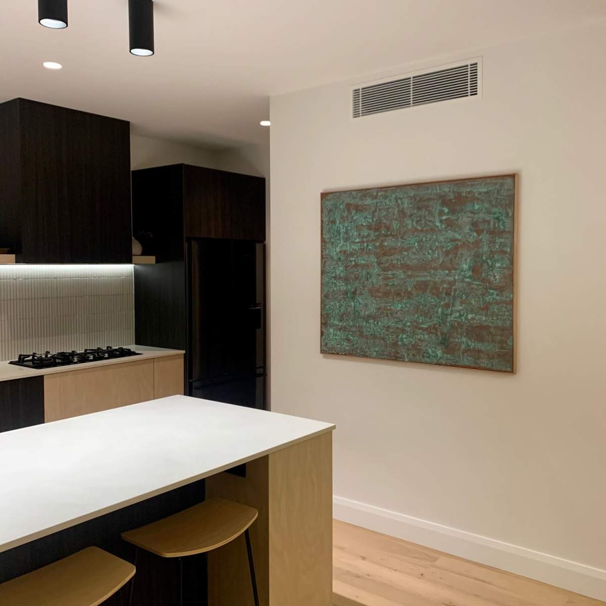 A modern kitchen features dark cabinetry, a white countertop, and a large textured wall art piece.