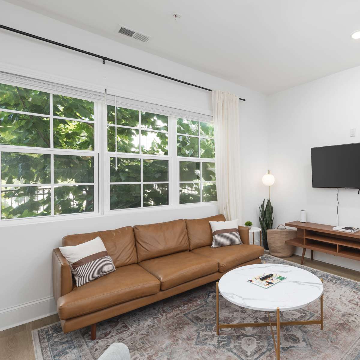 A modern living room with a brown leather sofa, a round coffee table, a wooden TV stand, and large windows allowing natural light to enter.