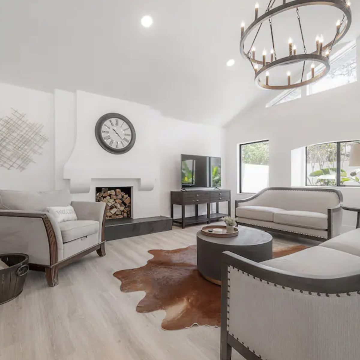 The living room features light-colored walls, a modern chandelier, a fireplace with stacked logs, and minimalist furnishings, including a circular coffee table and two sofas.