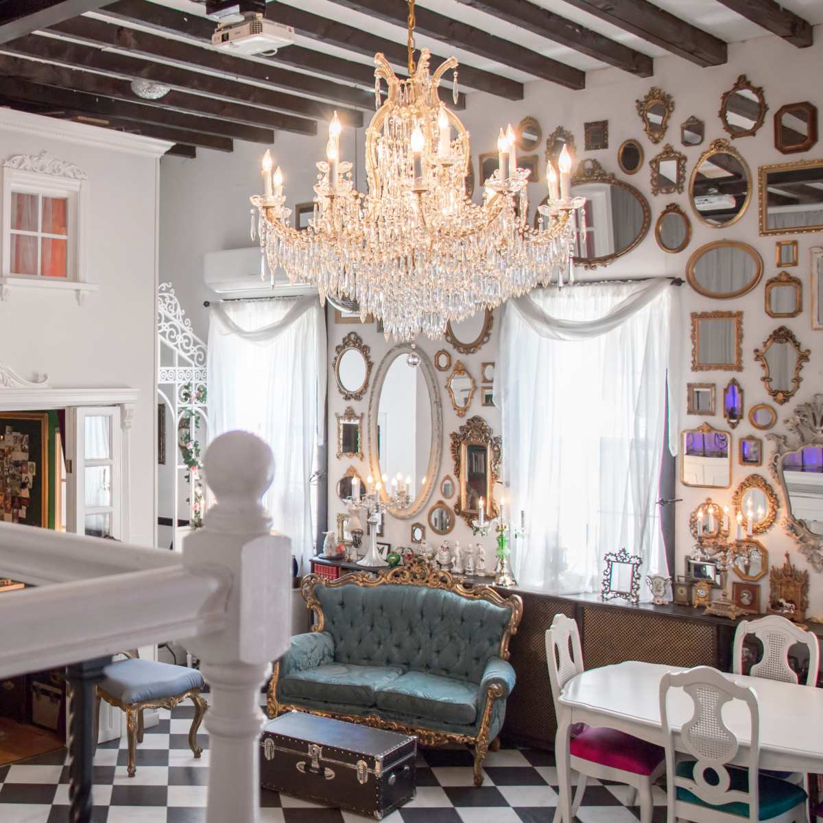 An elegantly designed interior features a vintage sofa, a crystal chandelier, and numerous framed mirrors on the walls, with a staircase leading to a small balcony.