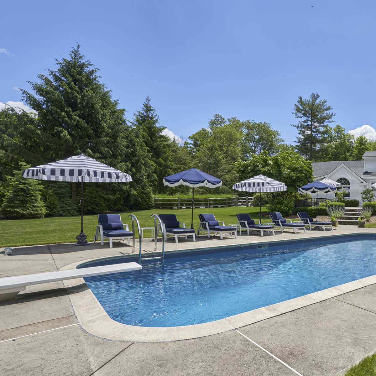 A residential backyard featuring a swimming pool surrounded by lounge chairs and several striped umbrellas, with a house and landscaped greenery in the background.