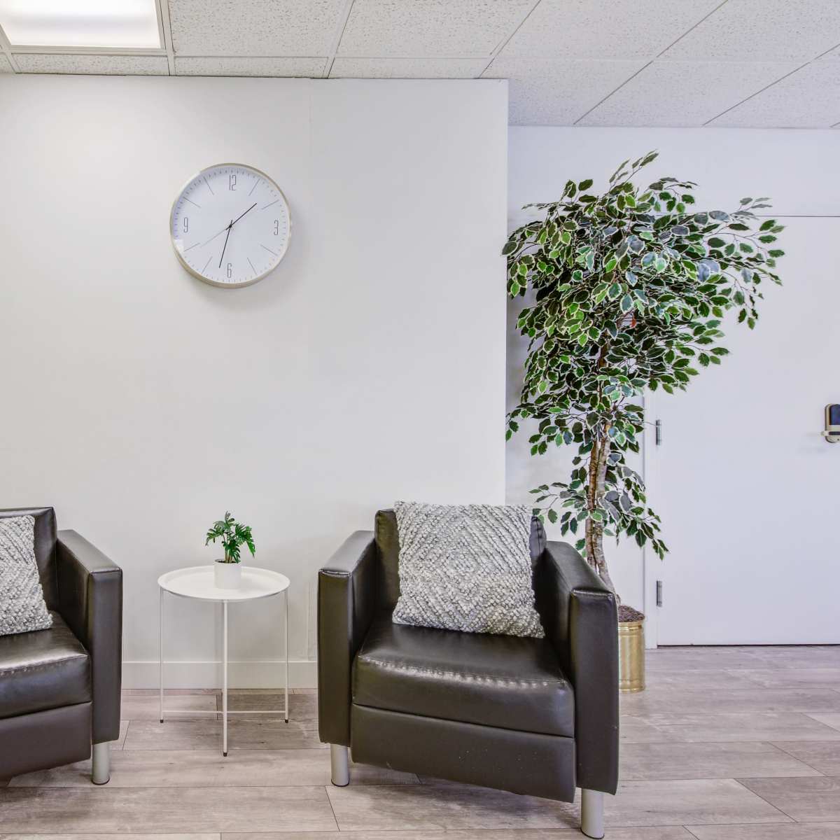 Two black armchairs with gray cushions positioned beside a small round table and a potted plant against a backdrop of a yellow wall and a white wall with a clock.