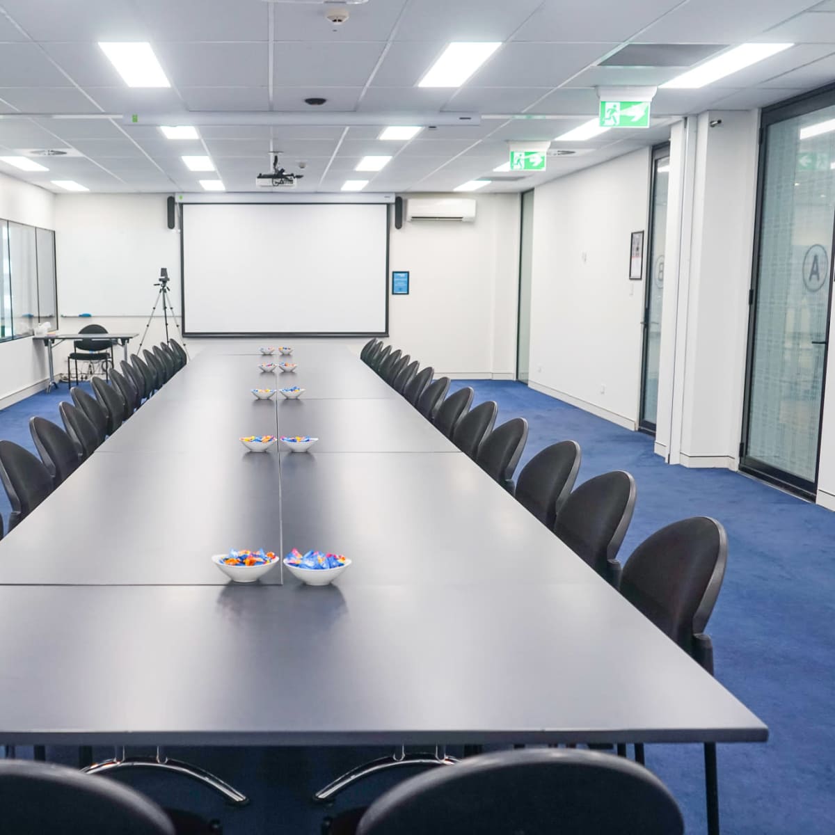 A long conference table with black chairs is set up in a modern meeting room, featuring bowls of snacks at each end and a projector screen at the front.