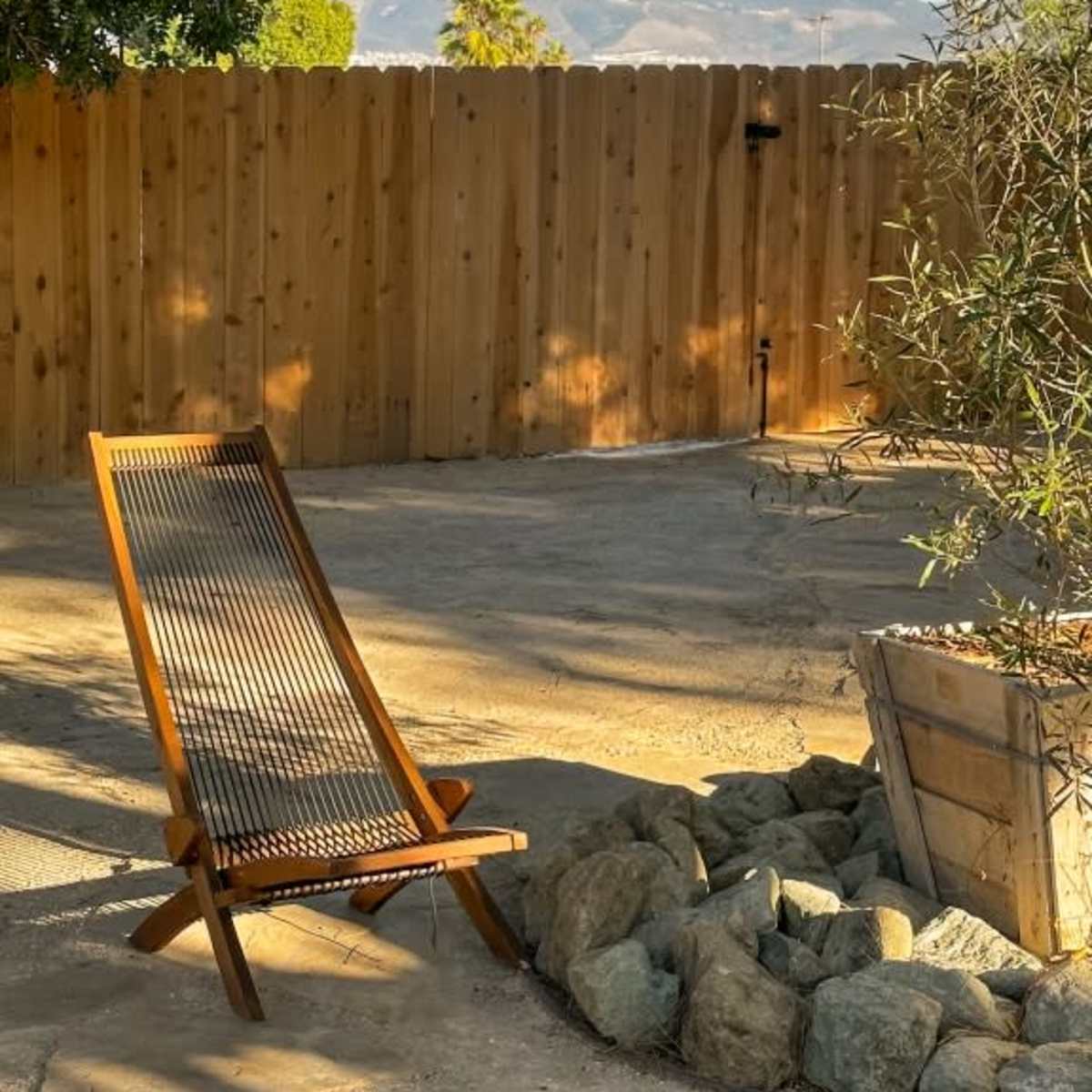 A wooden lounge chair is positioned next to a stone fire pit, surrounded by a gravel area and backed by a wooden fence and distant mountains.