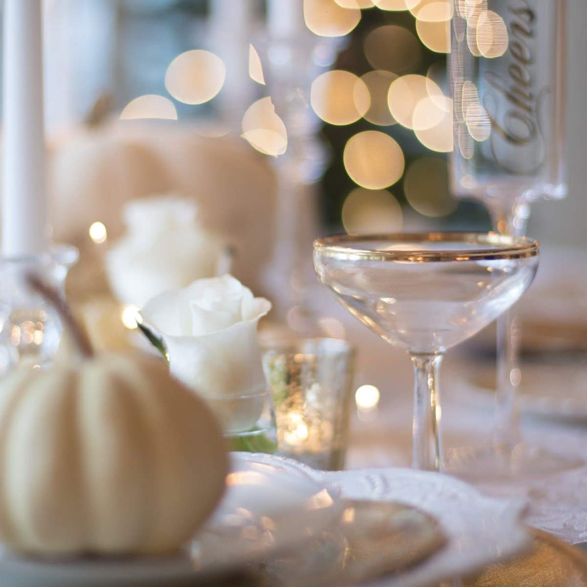 A elegantly set dining table features white pumpkins, roses, and sparkling glasses beside soft, blurred lights in the background.