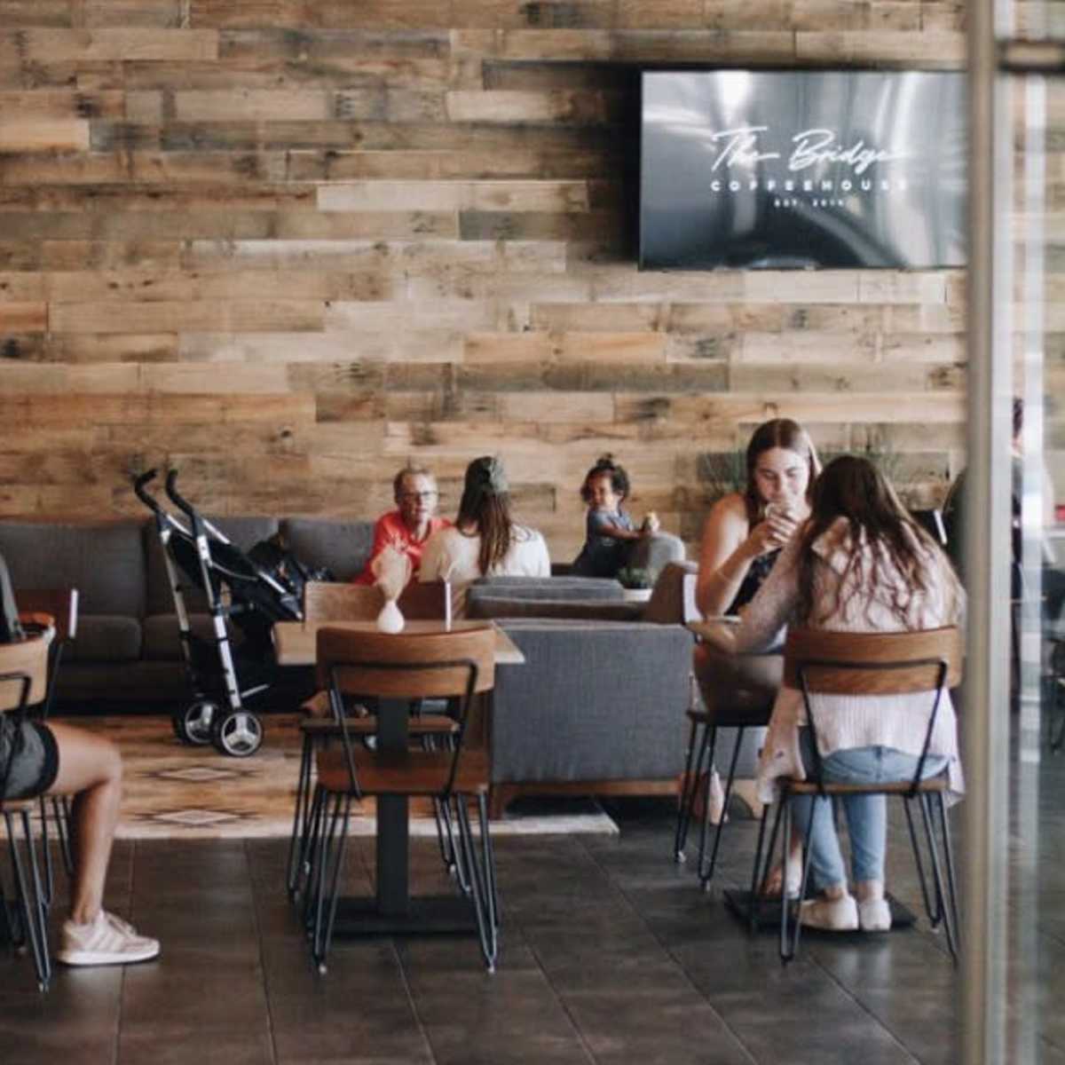 A modern coffee shop with wooden walls, where several people are seated at tables, some working on laptops, while a stroller is visible nearby.