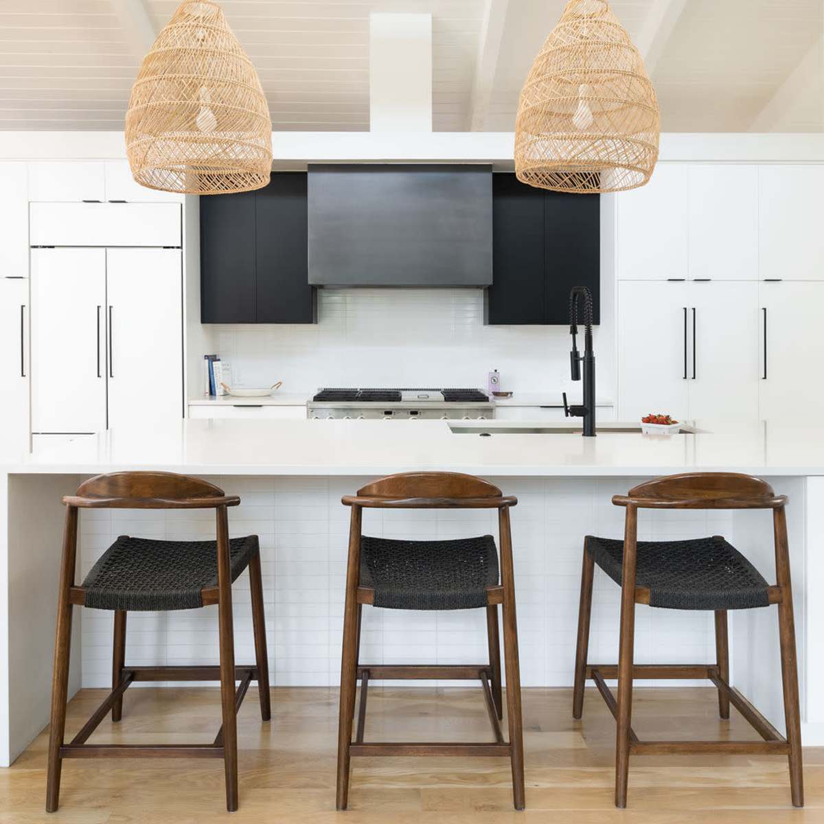 A modern kitchen features a central island with three wooden stools, white cabinetry, and two woven pendant lights hanging overhead.