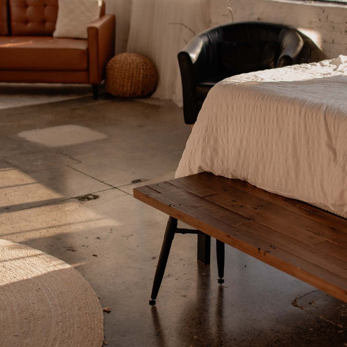 A modern interior of a room featuring a bed with a textured white cover, a wooden bench, a circular rug, a black chair, and a brown sofa beside large windows with sheer curtains.