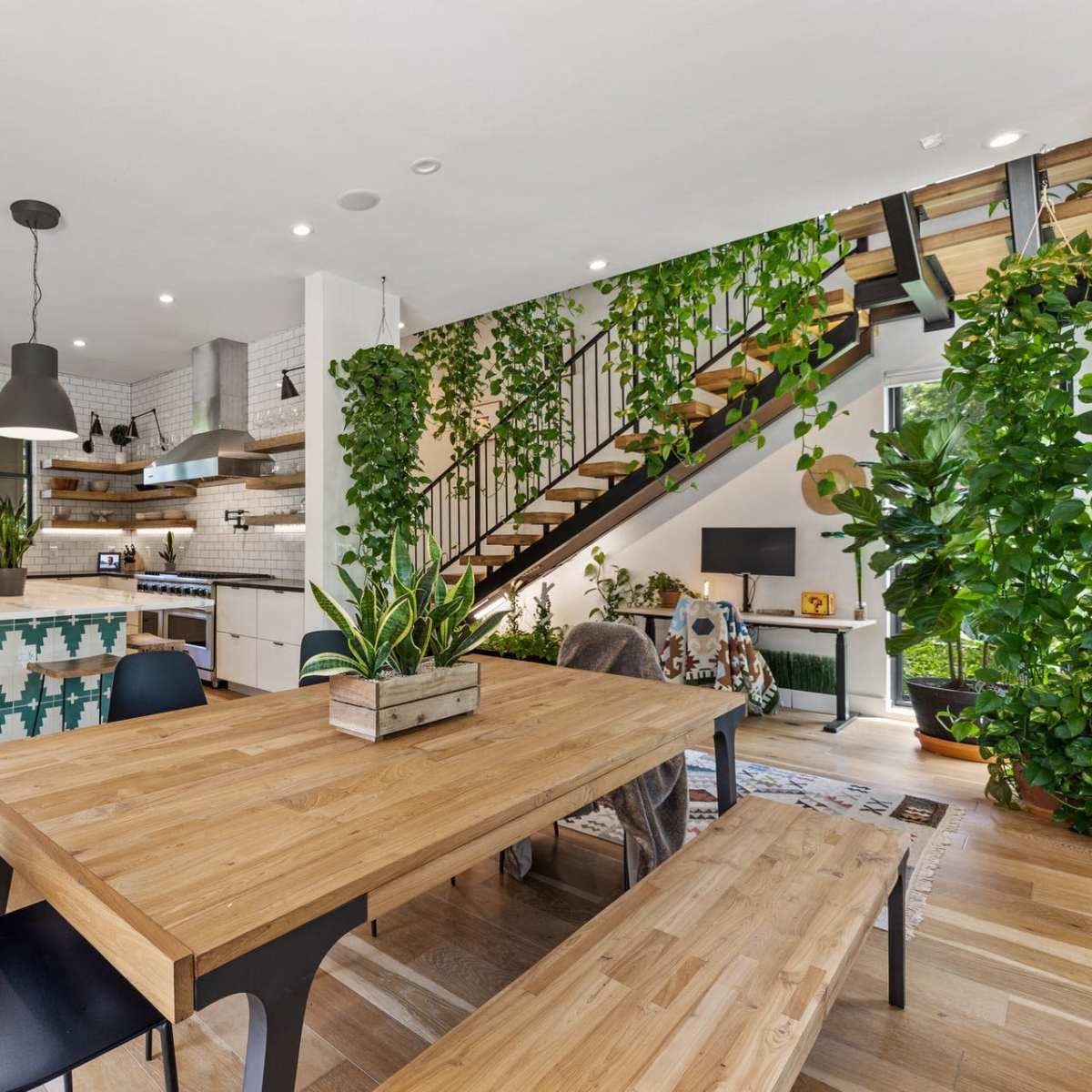 A modern kitchen and dining area features a wooden table, black chairs, and numerous plants, with a staircase in the background.