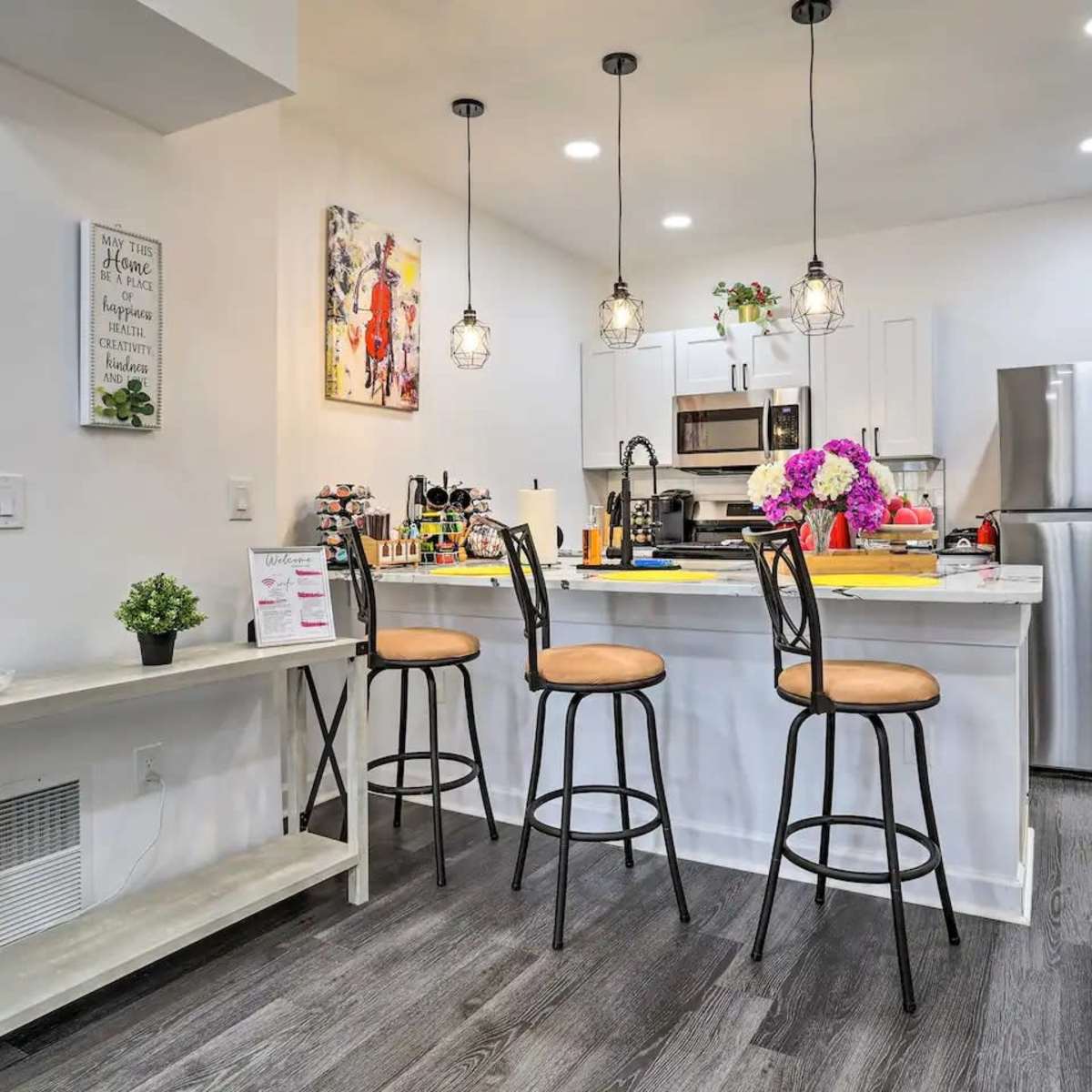 A modern kitchen with a breakfast bar featuring three stools, a stainless steel refrigerator, and decorative elements like flowers and artwork on the walls.