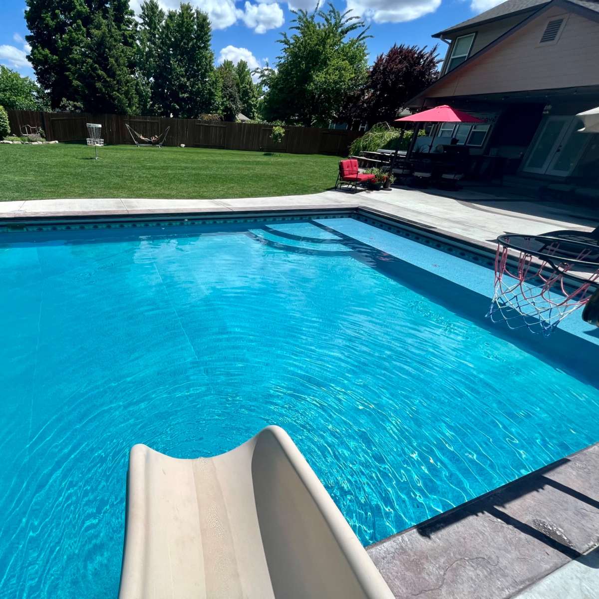 A clear swimming pool with a slide, surrounded by green grass and trees, and a patio area with seating and an umbrella.