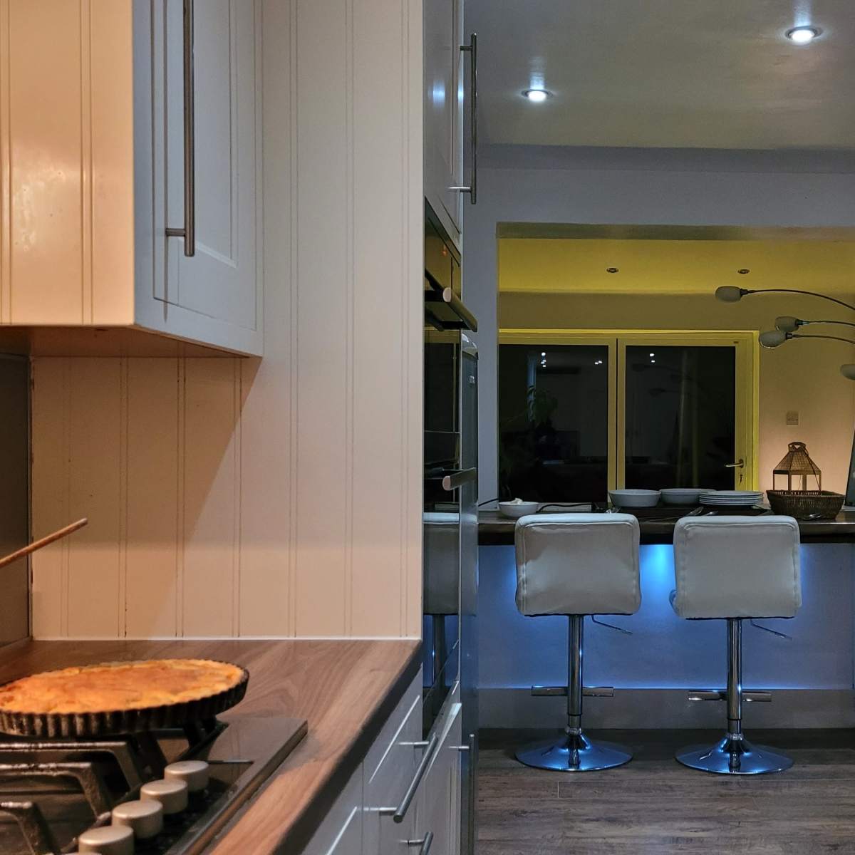 A modern kitchen features a pot on the stove and a bar area with two white stools, illuminated by soft lighting.
