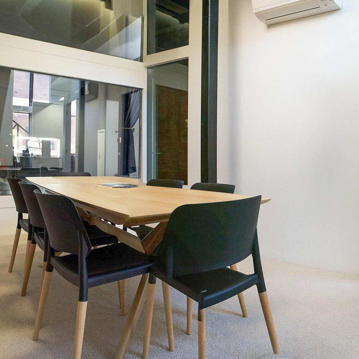 A minimalist conference room features a wooden table surrounded by black chairs, with a view into a brightly lit kitchen area through large glass panels.