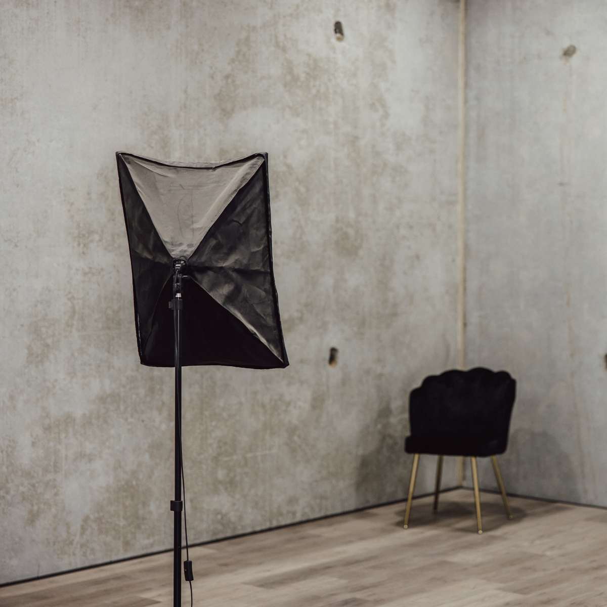 An empty room with a concrete wall, a black photography softbox on a stand, and a black chair with gold legs.
