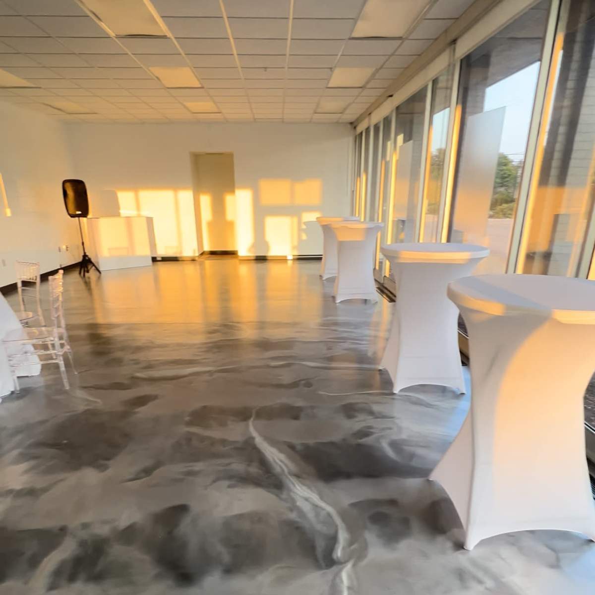 A modern event space with glossy, marbled flooring and several tall tables arranged along the windows, lit by warm afternoon sunlight.