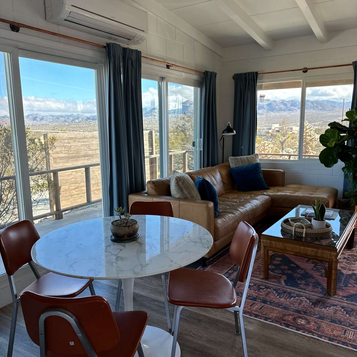 A living area with a circular marble dining table, a brown leather sofa, and large windows offering a view of a landscape with mountains in the background.