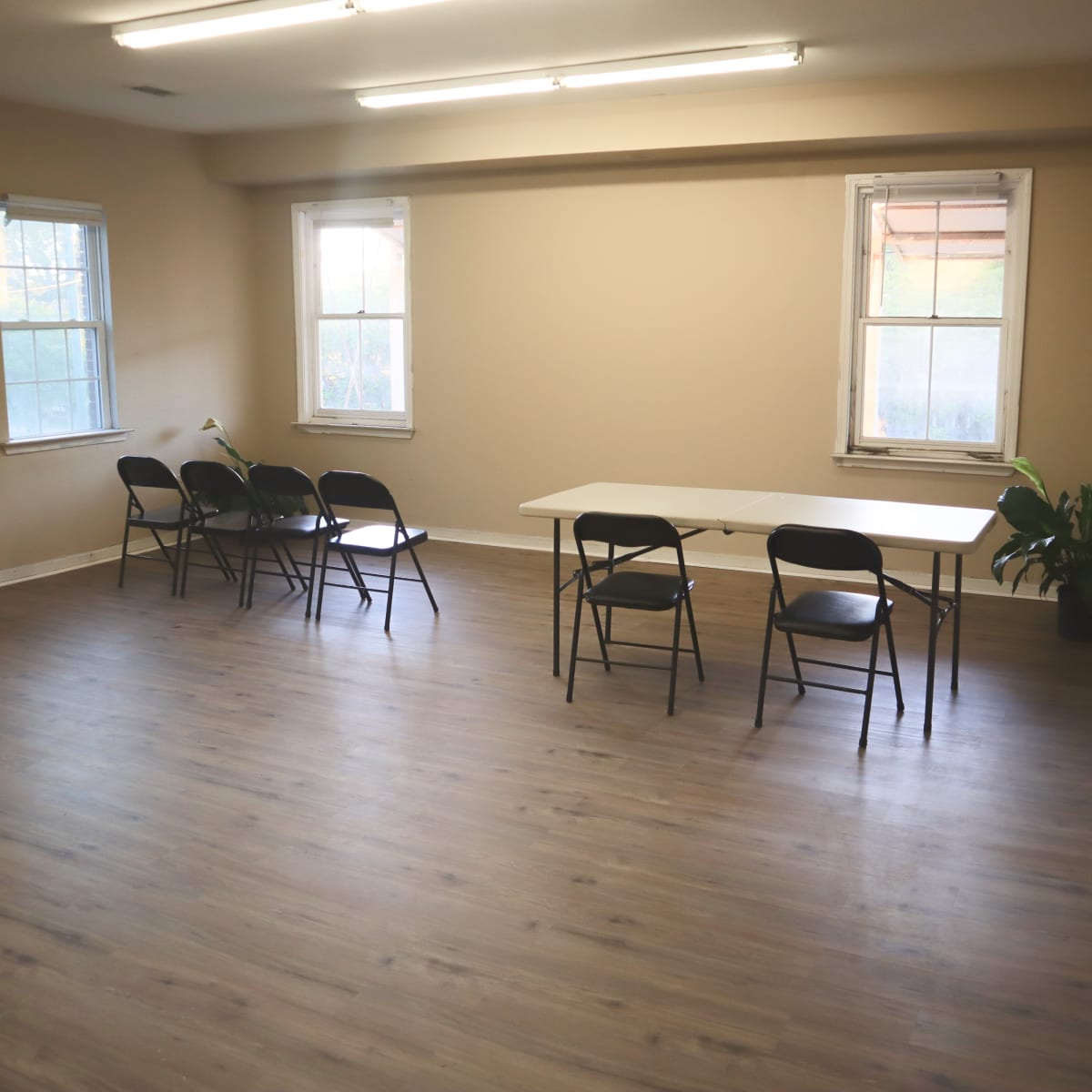 An empty room with a table and six black chairs arranged around it, and two windows allowing natural light.