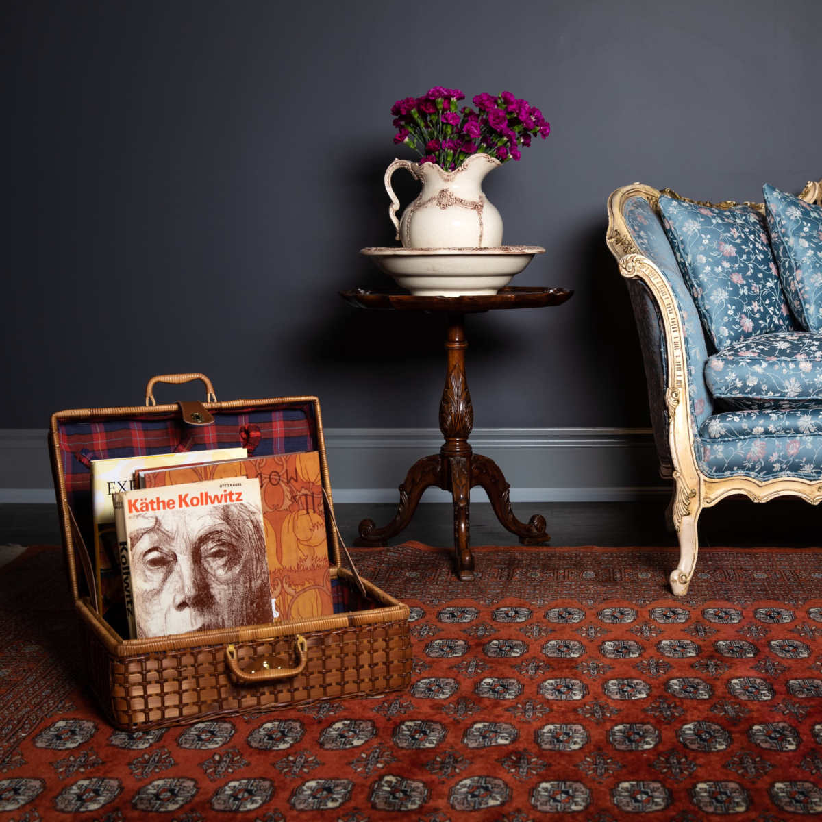 A blue floral-patterned sofa sits next to a side table with a vase of flowers, while an open suitcase on the floor displays a book.