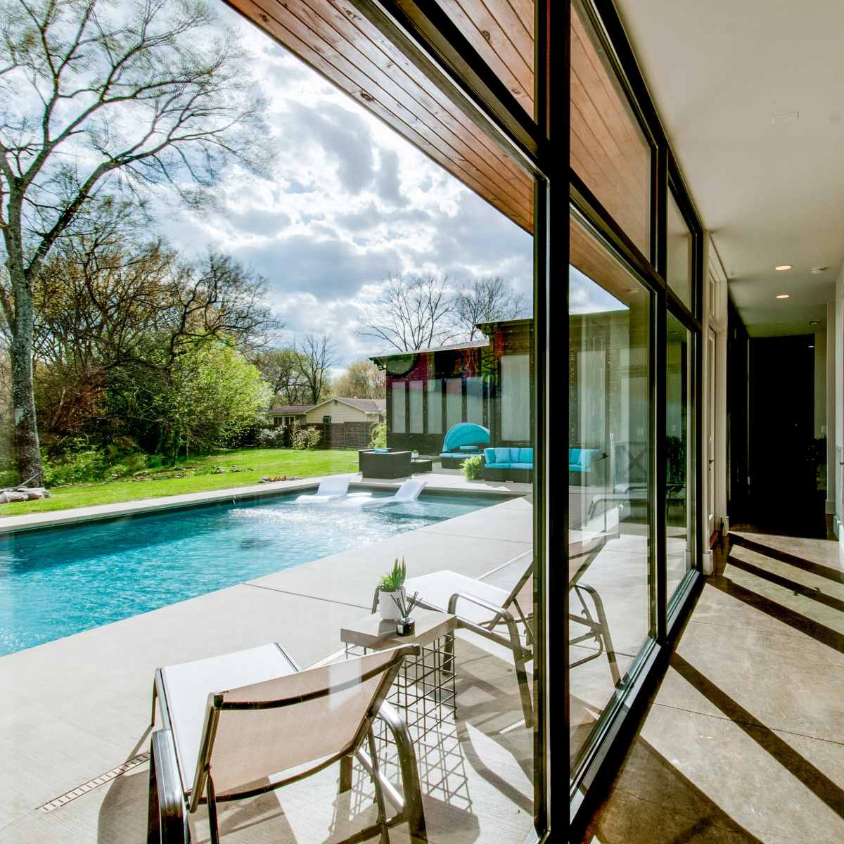 A modern home features large glass doors opening to a poolside area with patio chairs, surrounded by greenery and clouds in the sky.