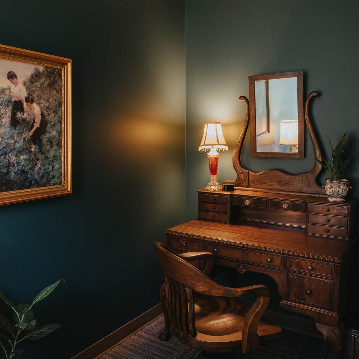 A corner of a room featuring a vintage wooden desk with a chair, an ornate mirror, a lamp, a potted plant, and a large framed painting of two girls in a garden.