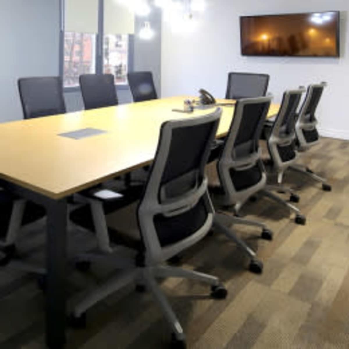 A long conference table surrounded by ergonomic chairs is set up in a well-lit meeting room with large windows and a wall-mounted screen.