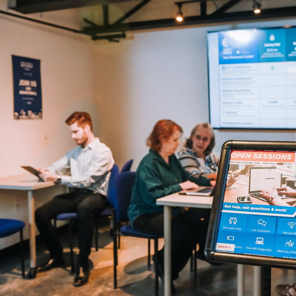 A group of people engaged in a discussion at a table in a room with a large display board behind them, while a digital sign promotes open sessions for asking questions.