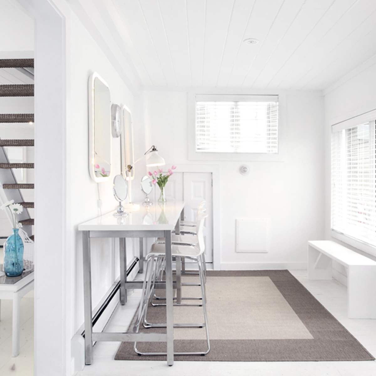 A bright, minimalist interior featuring a dining area with a table and chairs, a staircase, and large windows with blinds.