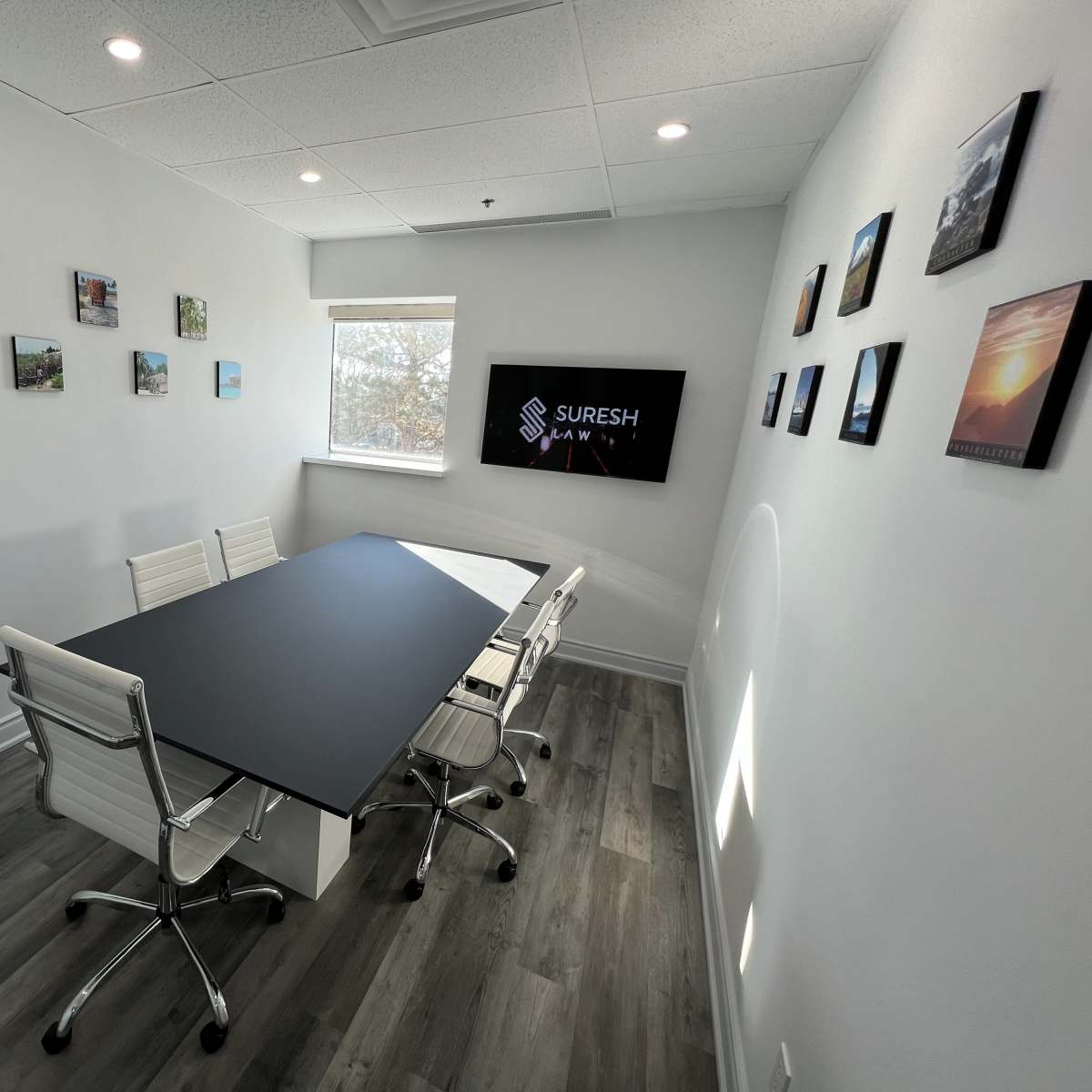 A modern conference room features a rectangular table surrounded by white chairs, with artwork on the walls and a display screen showing the name "Suresh."