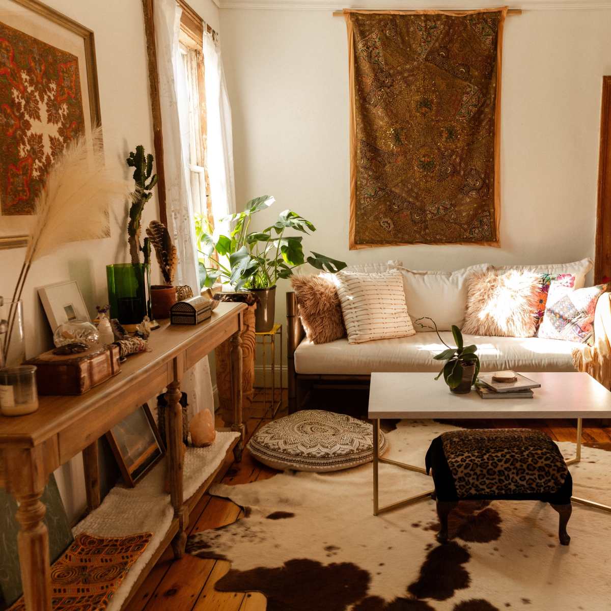 The room features a cozy living space with a white sofa, a wooden coffee table, and decorative elements including plants and a patterned wall hanging.