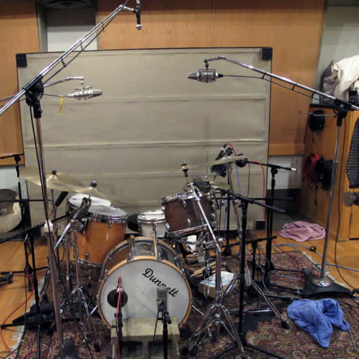 A drum set in a recording studio, surrounded by multiple microphones on stands and a soundproof backdrop.