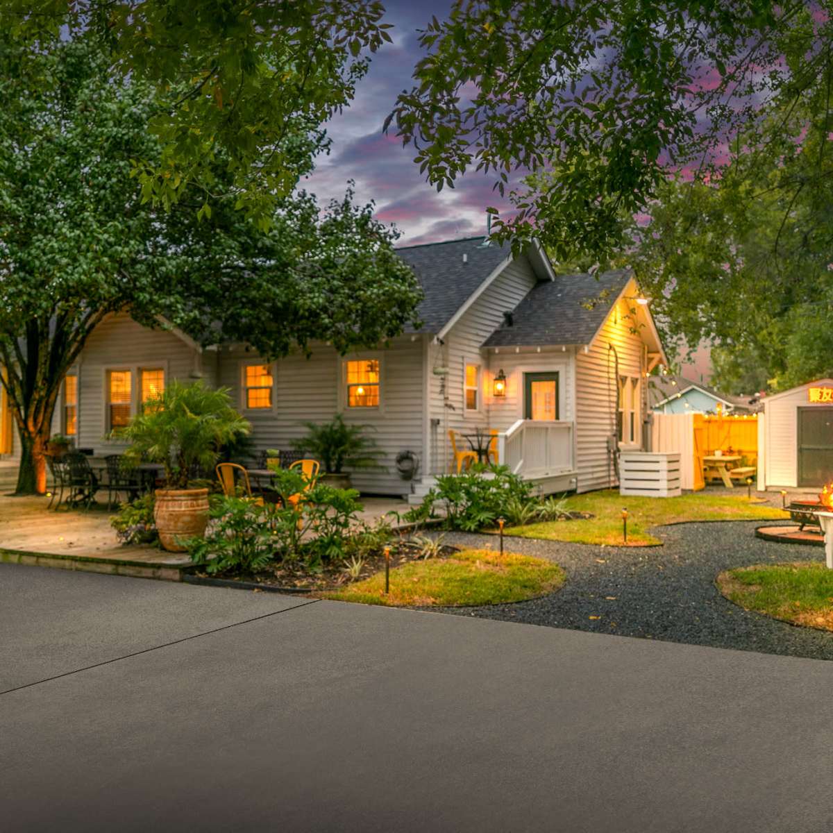 A well-lit backyard with a wooden house, outdoor seating, and a fire pit surrounded by lush greenery and garden lights at dusk.