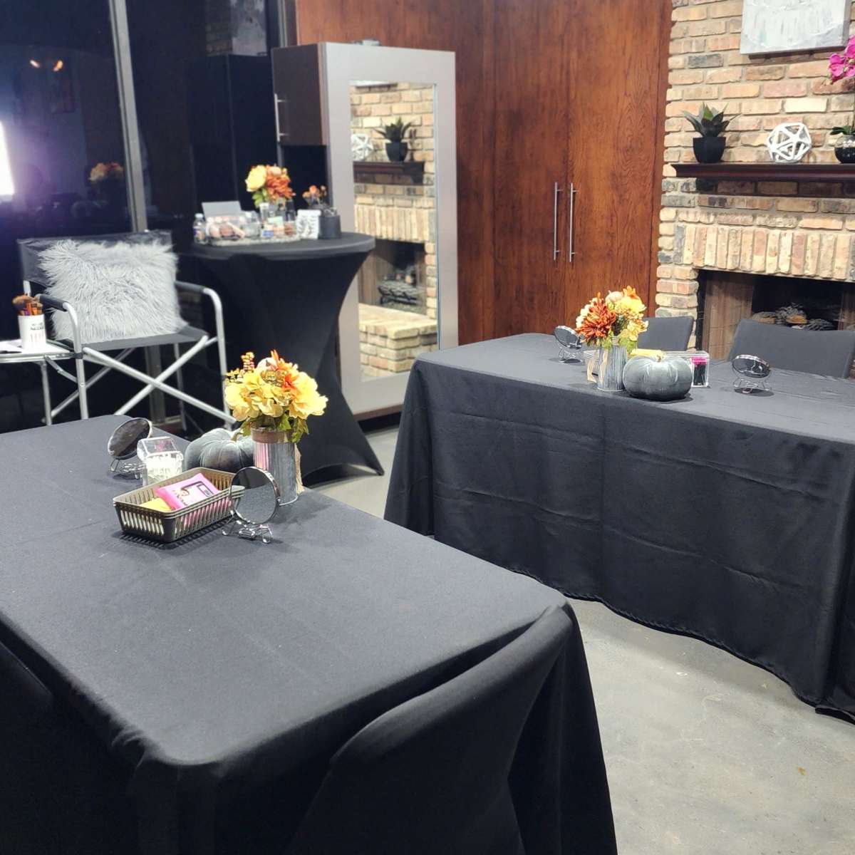 The scene features three tables with black tablecloths, adorned with floral arrangements, set up in a room with a brick fireplace and mirrored cabinets.