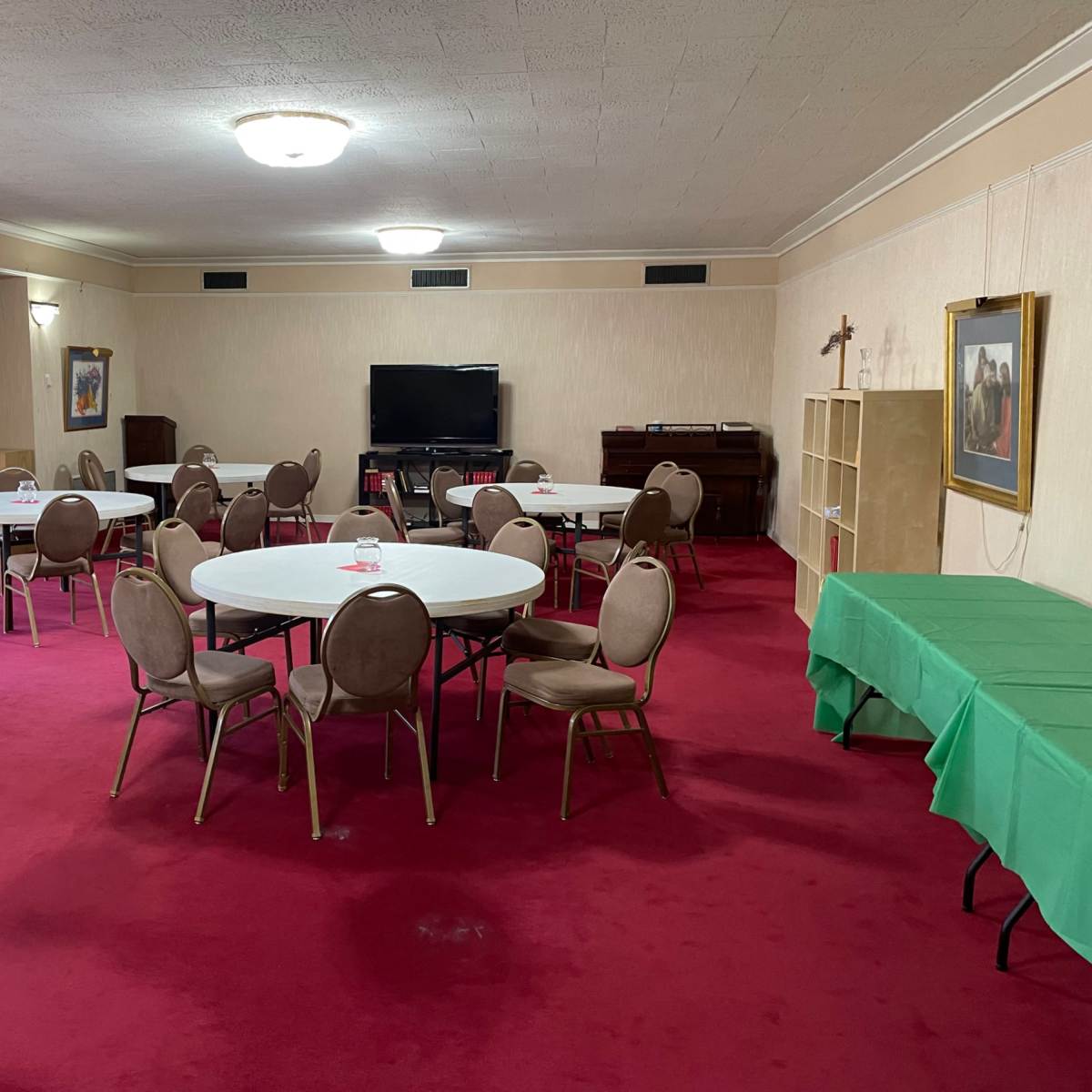 A spacious room with a red carpet, featuring several round tables surrounded by chairs, a television, a piano, and a green table along one wall.