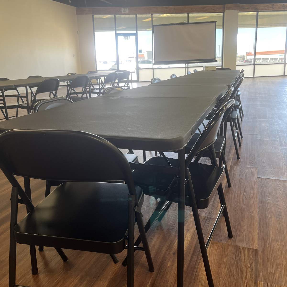 A spacious room with multiple rows of black folding tables and chairs, facing a large projector screen.